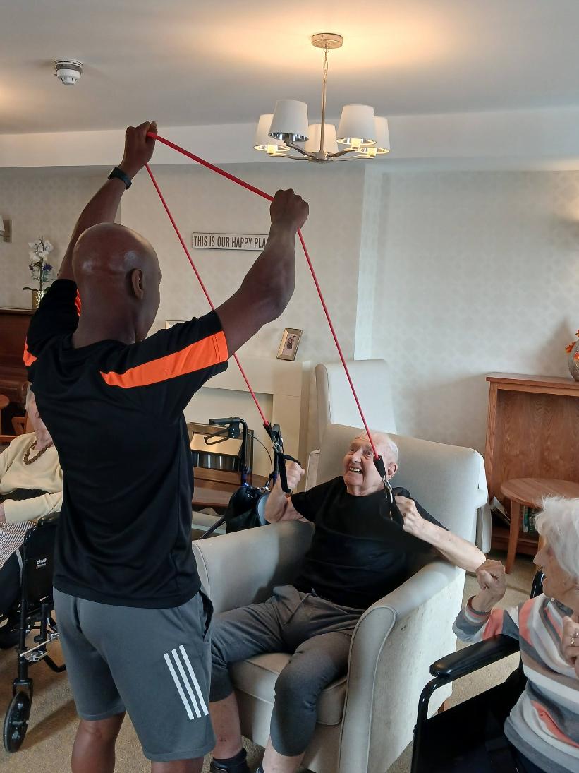 man helping an older man sat in a. chair exercise by lifting an exercise cable