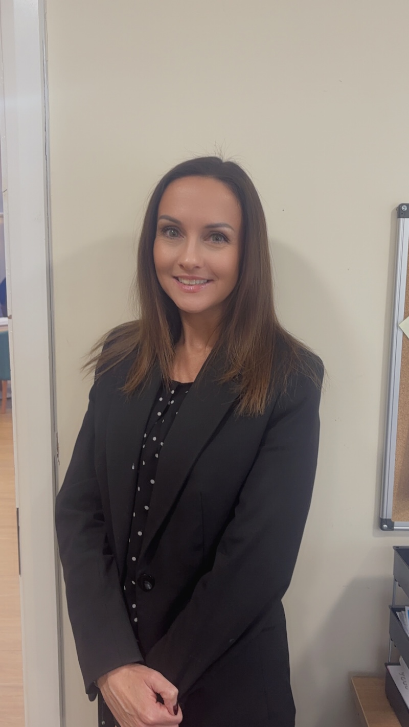 woman with brown hair wearing a black blazer stood smiling having her headshot taken for the team page
