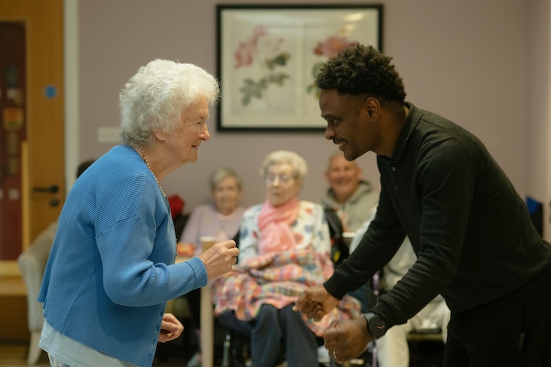 carer wearing a black jumper bend down smiling and dancing with a female resident wearing a blue jumper